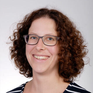 Portrait einer Frau mit lockigem Haar und Brille, vor neutralem Hintergrund.
